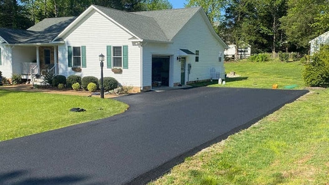 A freshly paved black asphalt driveway leading to a white house with a green lawn and shrubs.