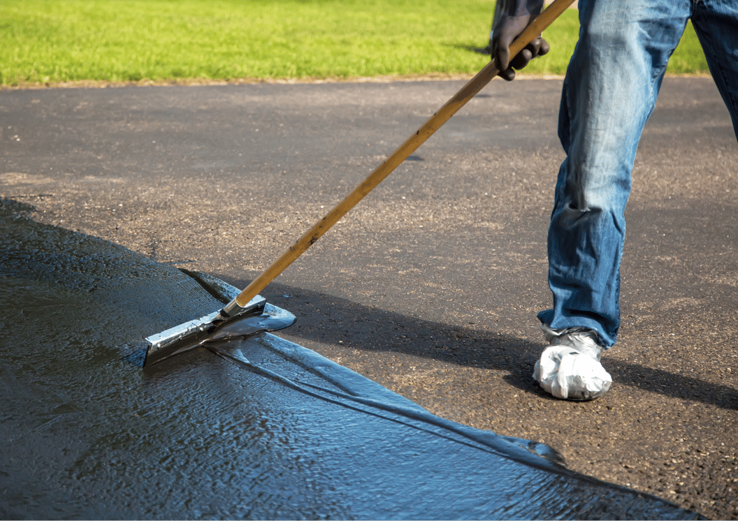 Person spreading black sealant on a driveway with a long-handled tool, wearing jeans and protective shoe covers.
