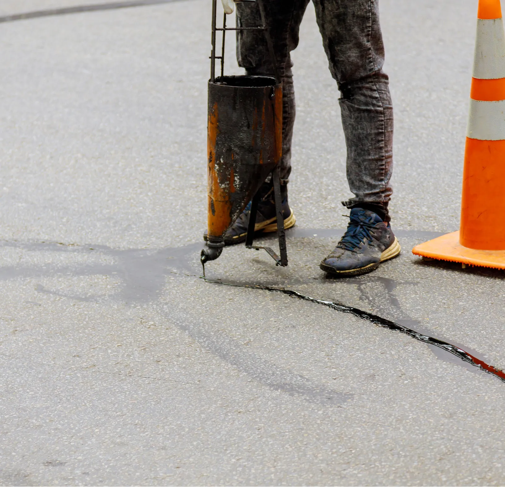 Worker sealing a crack in the road with a hot sealant, orange cone nearby on asphalt.