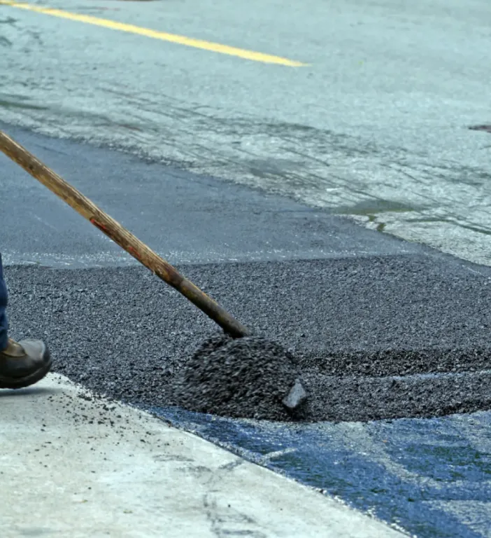 A worker spreads fresh asphalt on a road with a shovel, repairing the pavement near a curb.