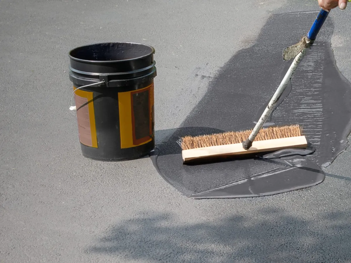 A person spreads black sealant on asphalt with a broom next to an open bucket.