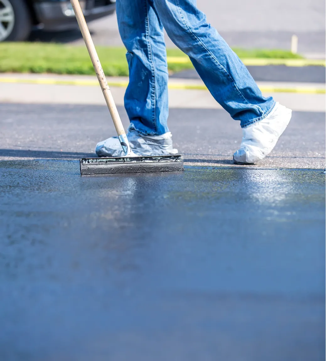 Person spreading sealant on asphalt pavement with a squeegee, wearing jeans and shoe covers.