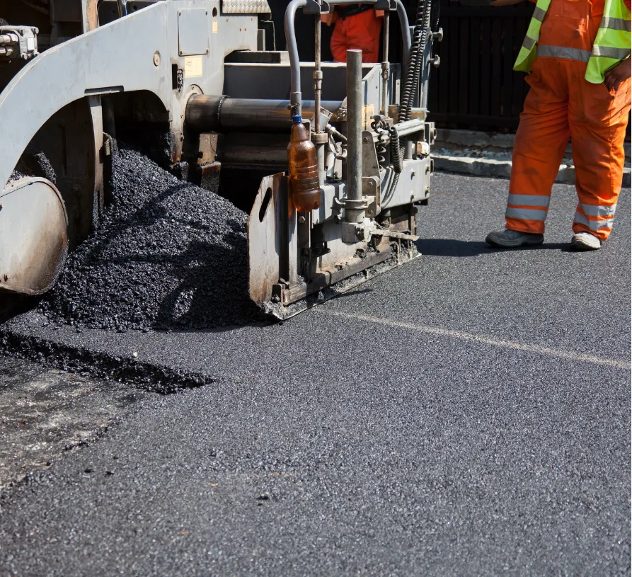 Asphalt paving machine laying new road with workers in orange safety uniforms nearby.