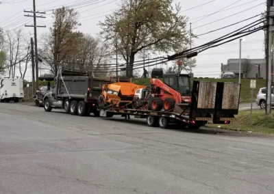 A flatbed trailer carries construction equipment, including a skid steer loader and orange machinery, on a road.