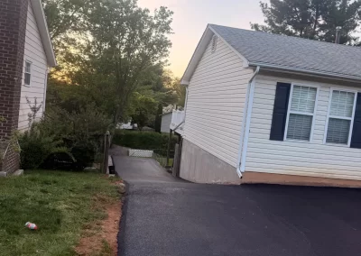 A paved driveway slopes down beside a white house with trees and a sunset in the background.