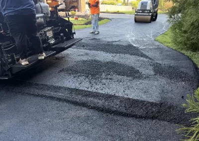 Workers paving a driveway with fresh asphalt using heavy machinery near a brick house.