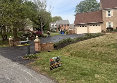 A brick driveway entrance and a yard sign on grass in front of suburban homes on a cloudy day.