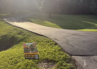 Sunlit driveway curves through grassy lawn with a yard sign advertising sealcoating services by the road.