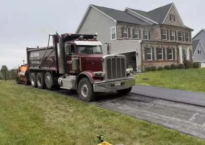 A dump truck and paver lay asphalt on a driveway in front of a two-story suburban house.