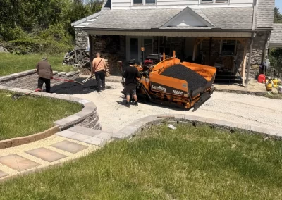 Three people work near an orange asphalt paver in a gravel driveway in front of a two-story house.