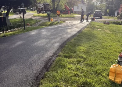 Workers paving a residential road with fresh asphalt on a sunny day, trees and houses nearby.