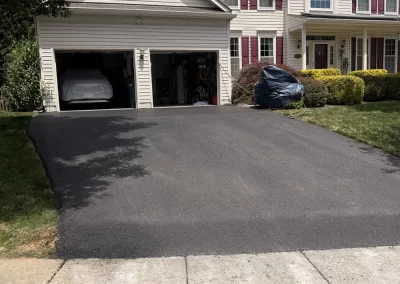A freshly paved driveway leads to a two-car garage in front of a beige suburban house with red shutters.