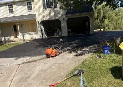 A house with fresh asphalt on the driveway, tools, and a "For Sale" sign on the grass nearby.