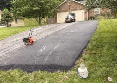 Freshly paved asphalt driveway with roller machine in front of a brick house, surrounded by green trees.