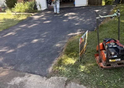 Worker in orange vest on newly paved driveway; garage open; paving machine and sign on grass nearby.