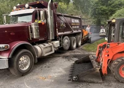 A dump truck and small loader are working on paving a road surrounded by green trees.