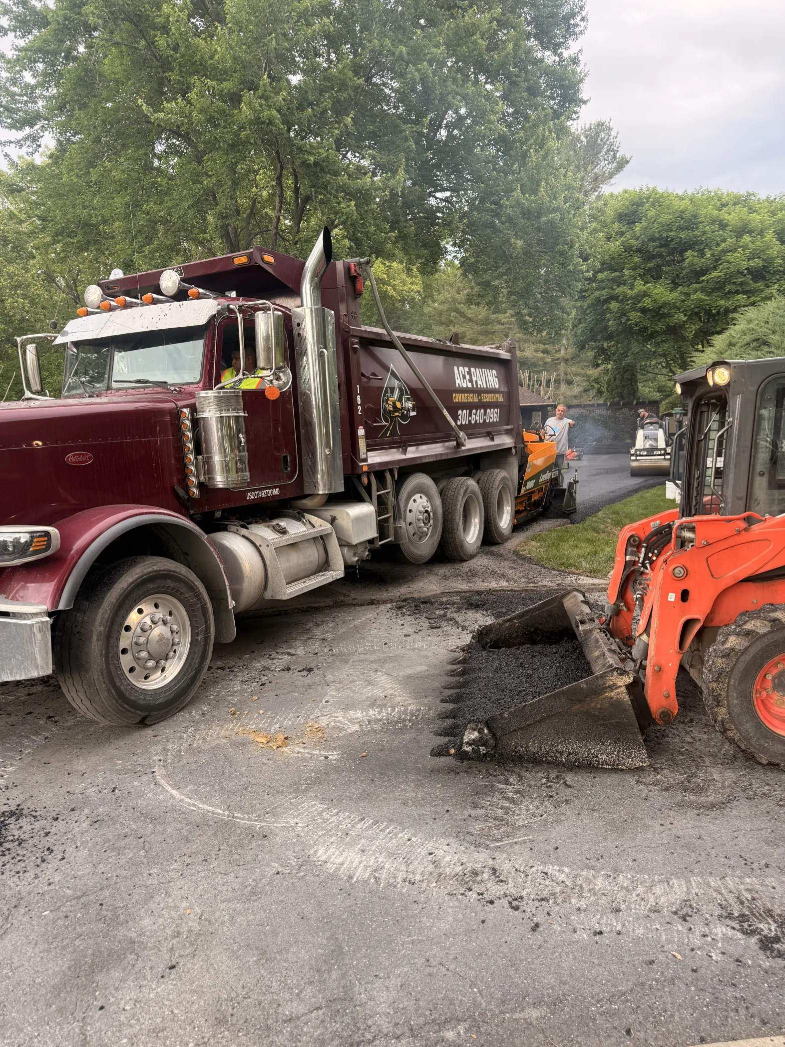 A dump truck and small loader are working on paving a road surrounded by green trees.